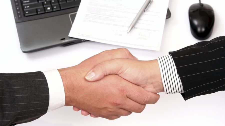 Two arms in pinstriped suits shaking hands with laptop, mouse, pen &amp; paper in background